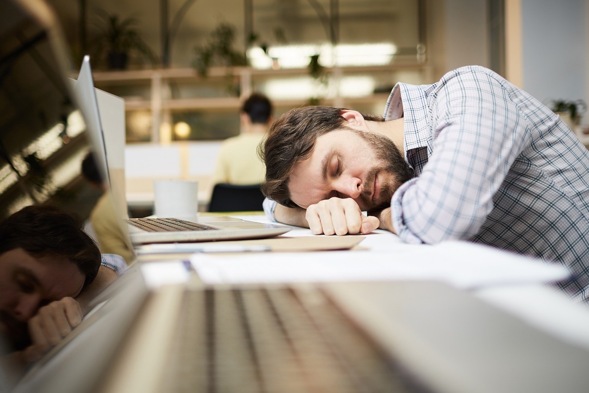 Homem cansado: employer branding Homem em escritório com ar de cansado debruçado sobre mesa de madeira com olhos fechados e notebooks à sua frente, além de papéis com dados impressos e colega de costas trabalhando ao fundo da imagem em desfoque