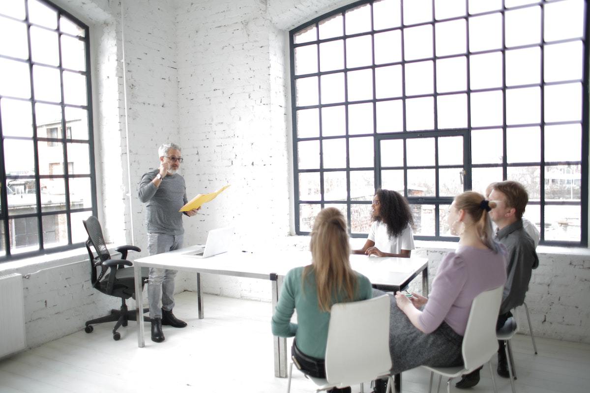 Reunião com time de marketing em ambiente rústico com pessoas sentadas diante de mesa e líder de pé explicando dados na frente de janela de vidro com grade de ferro em prédio
