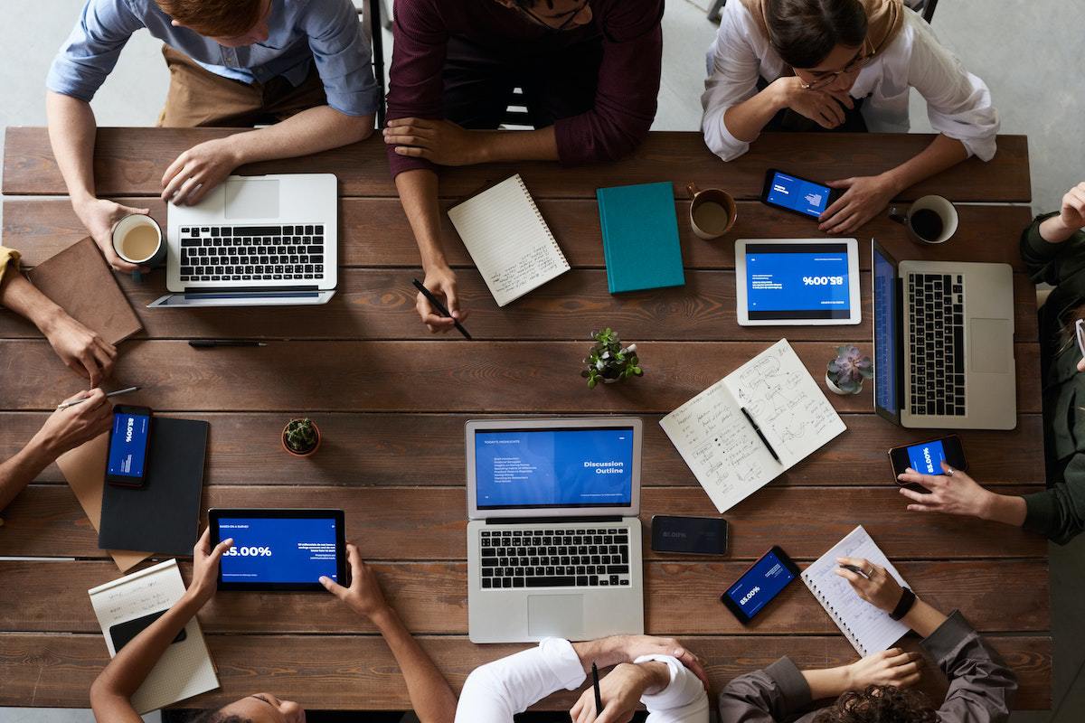 Equipe de marketing sentada ao redor de mesa de madeira retangular em reunião analisando dados impressos e em tablets e notebooks à frente de cada um deles