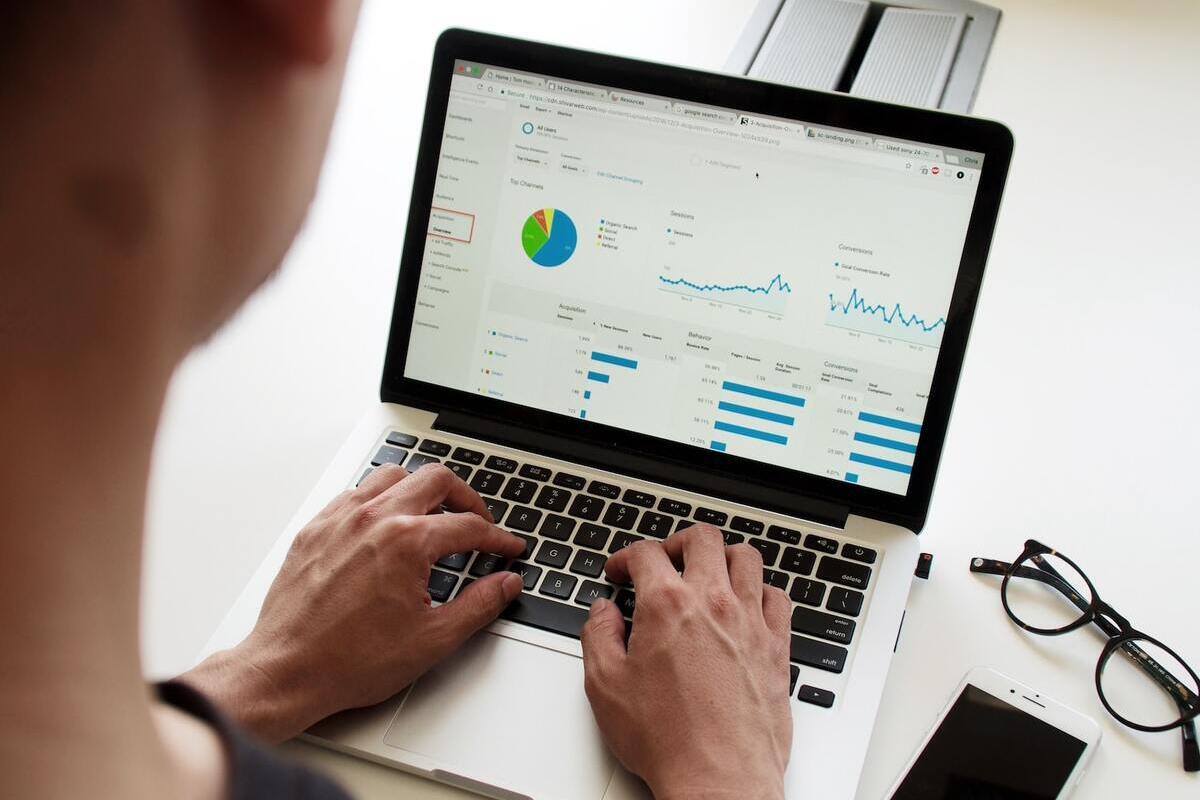 Homem sentado diante de notebook analisando resultados de números com vários gráficos e métricas apoiado sobre mesa branca com seu smartphone e seu óculos à direita