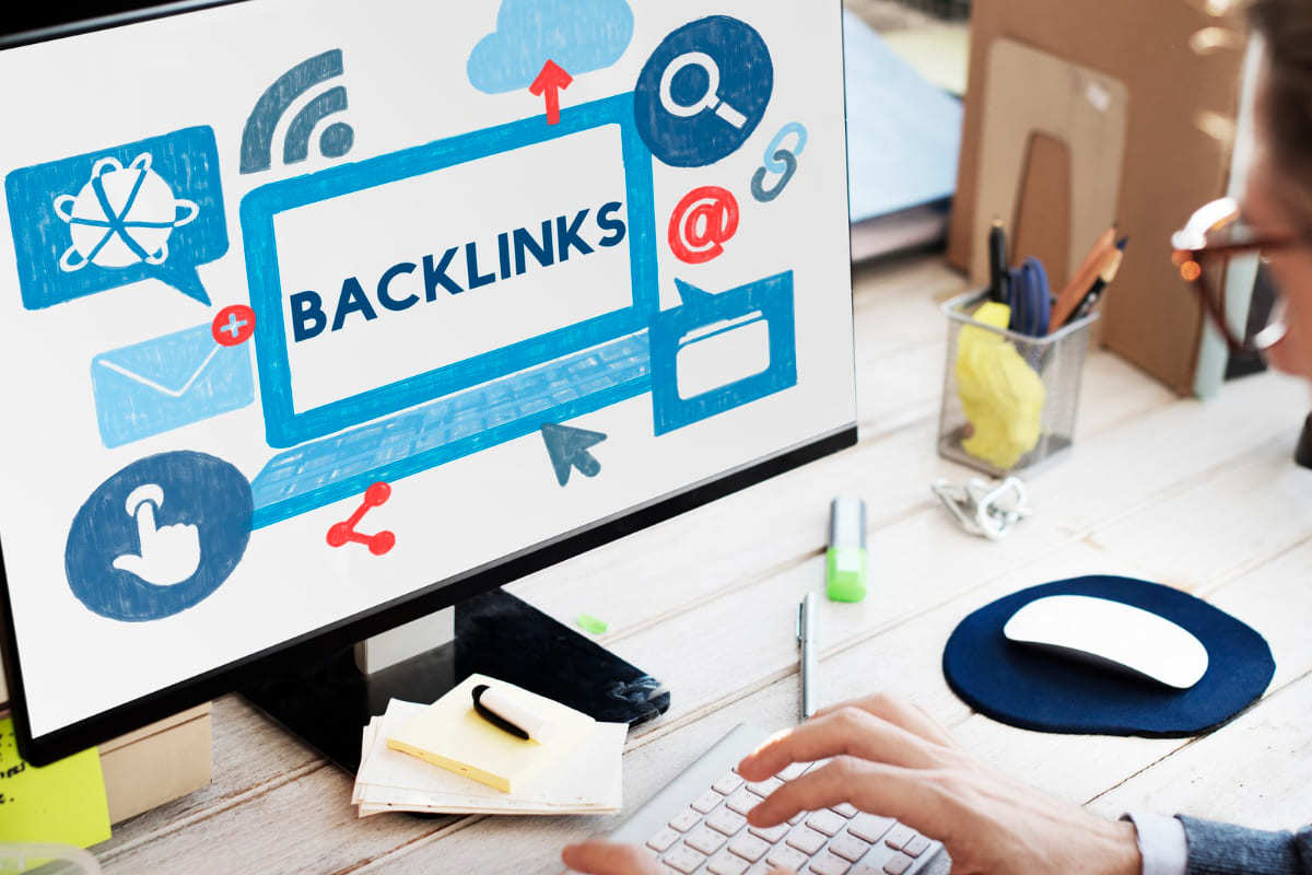 Pessoa sentada diante de computador sobre mesa e usando o mesmo, que está com montagem sobre o tema backlinks na tela e tem objetos como porta-canetas e post-its espalhados ao seu redor