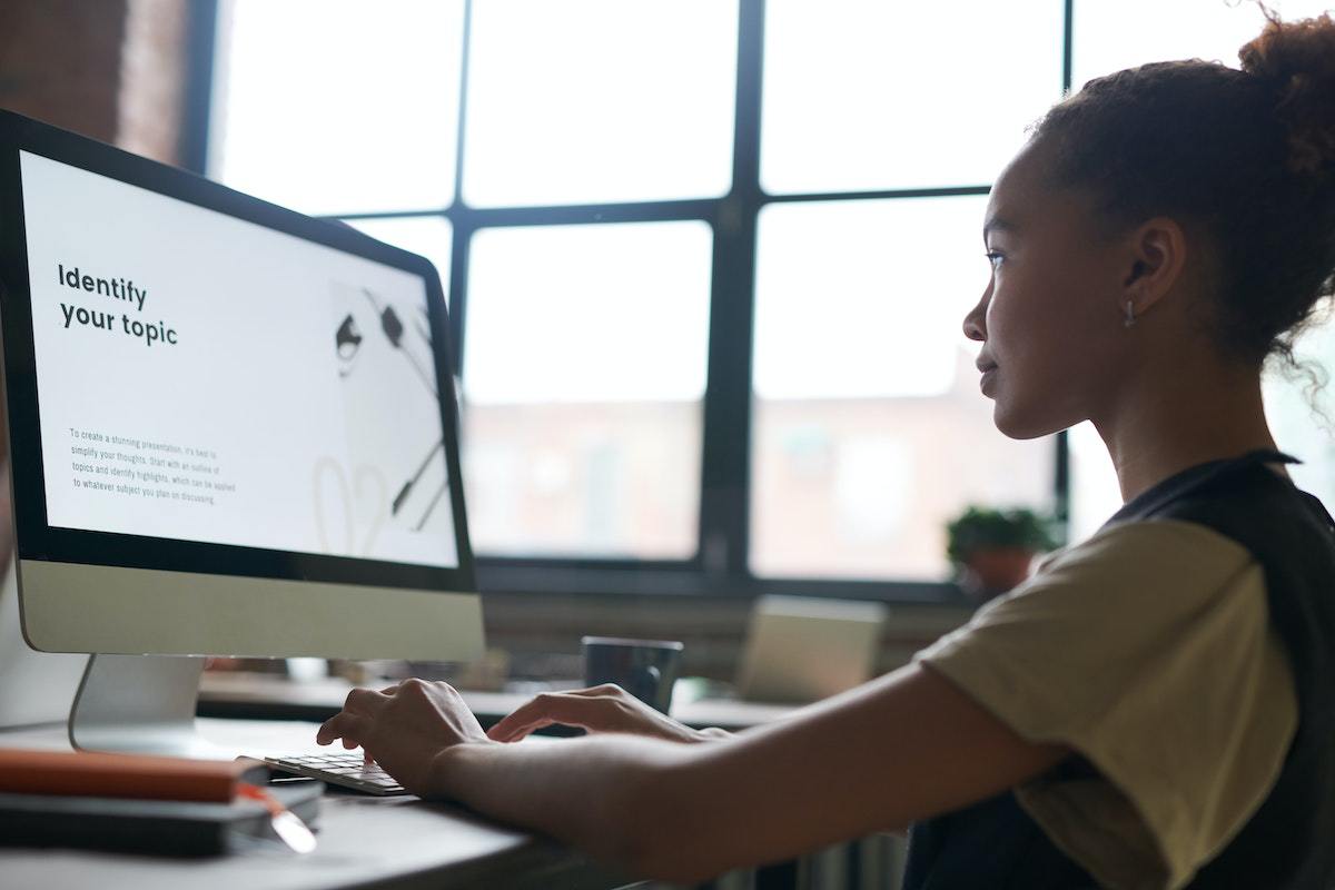 Mulher trabalhando em escritório diante de janela de vidro usando computador branco sobre mesa de madeira com montagem com frase Identify your topic