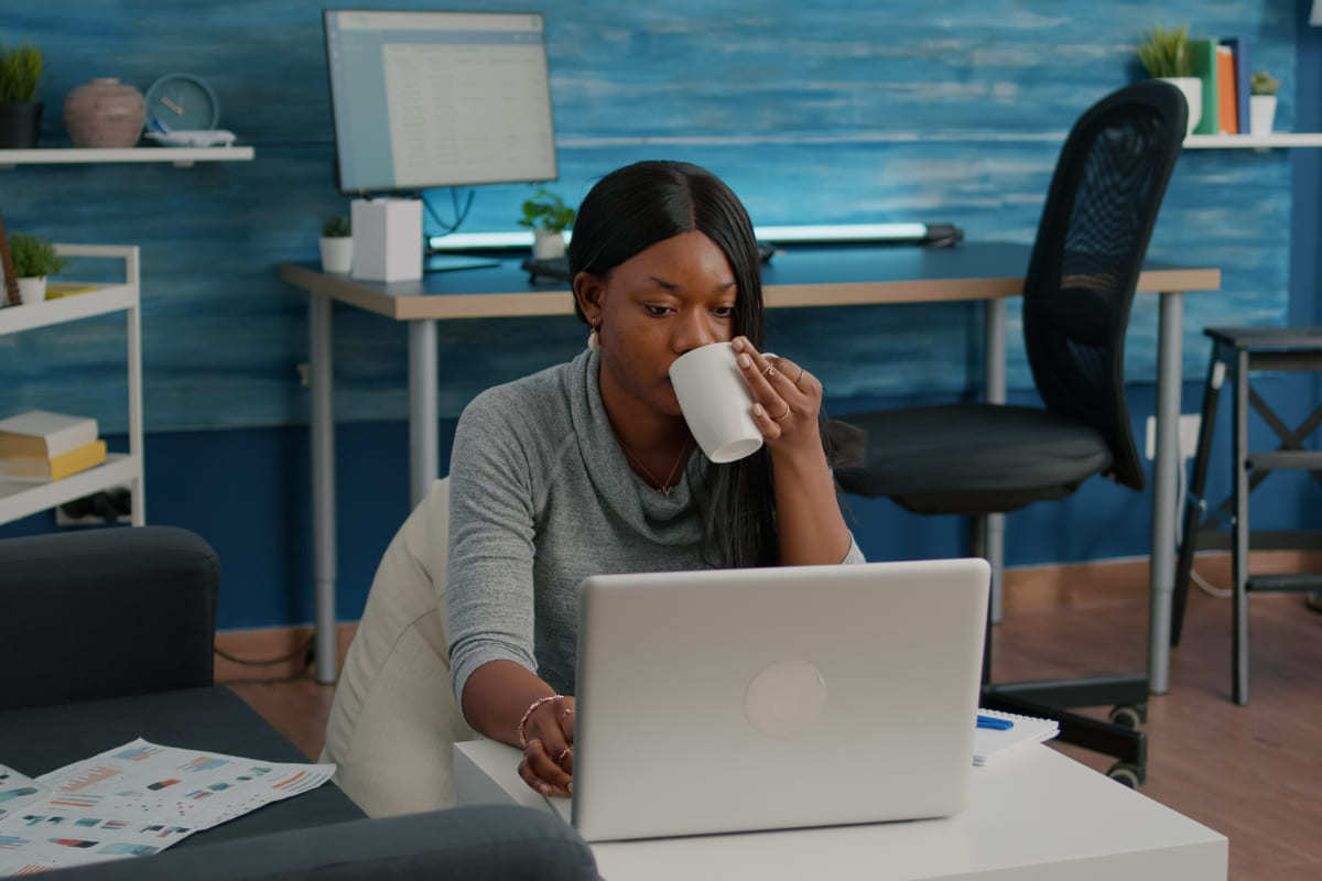 Mulher trabalhando Mulher tomando bebida em caneca branca usando notebook sobre mesa branca retangular com objetos de estudo e trabalho ao fundo da imagem