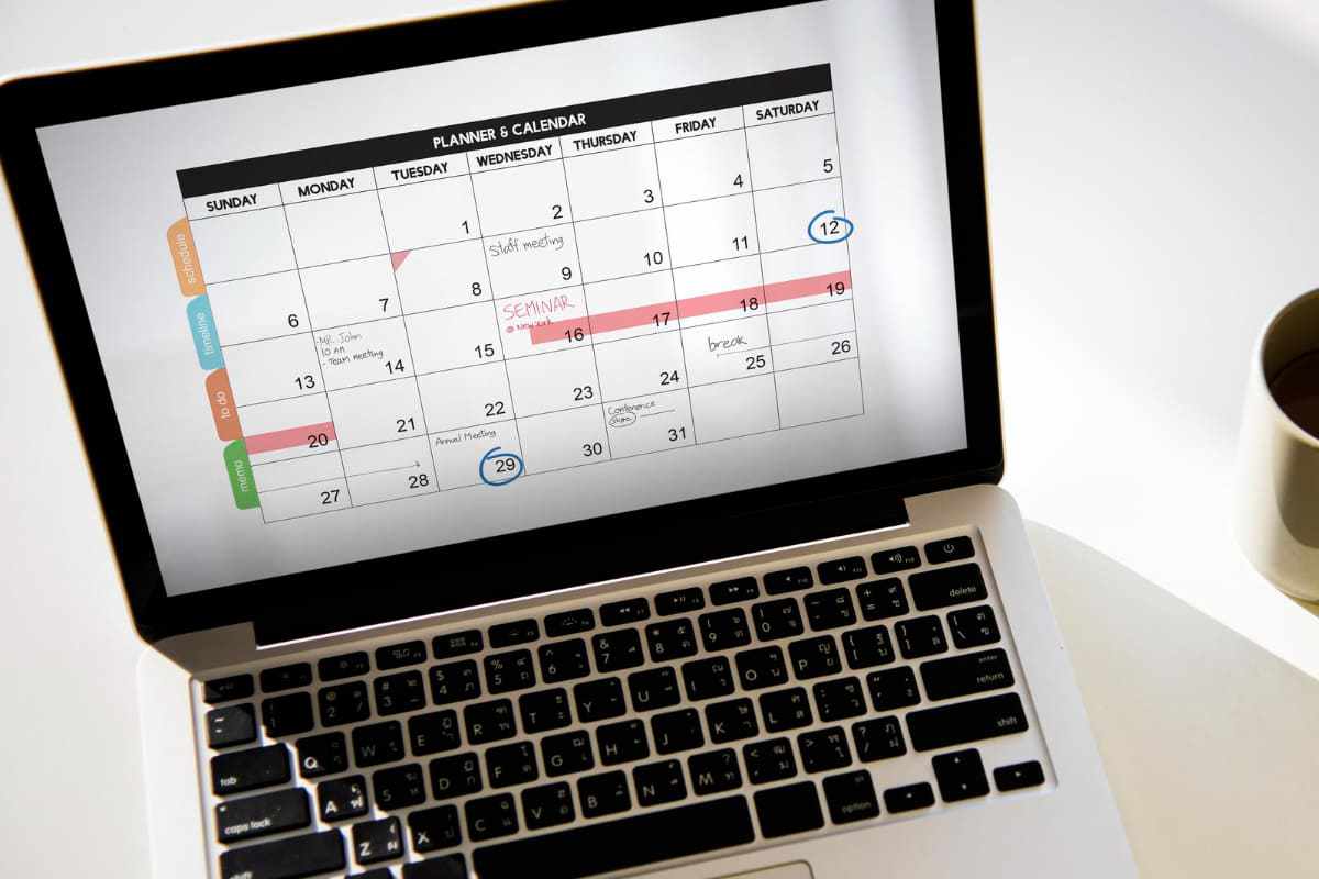 Notebook com agenda: Frequência de postagem Notebook sobre mesa e ao lado de copo com agenda na tela em parte do calendário