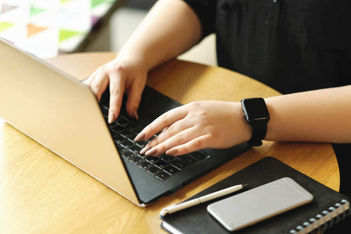 Notebook: Frequência de postagem Mulher sentada diante de mesa pequena de madeira redonda usando notebook e com agenda, celular e caneta à sua esquerda