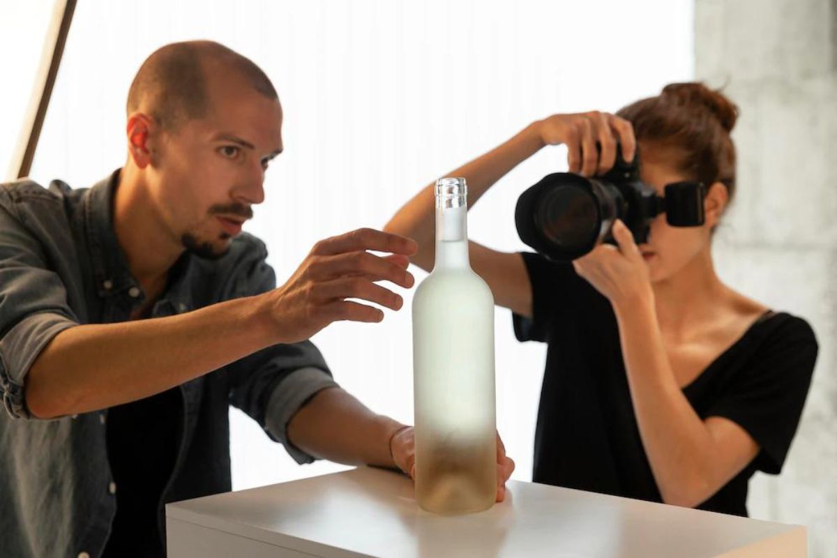 Homem segurando garrafa de vidro sobre pequena superfície branca e mulher fotografando o produto