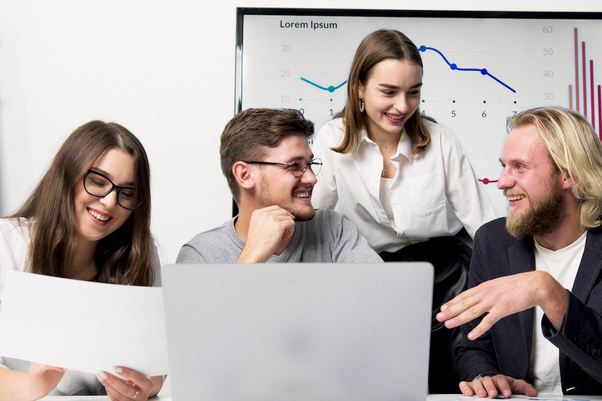 Local de trabalho: importância do marketing pessoal Dois homens e duas mulheres em local de trabalho conversando sobre algum tema enquanto sorriem diante de notebook com folhas de papel em suas mãos
