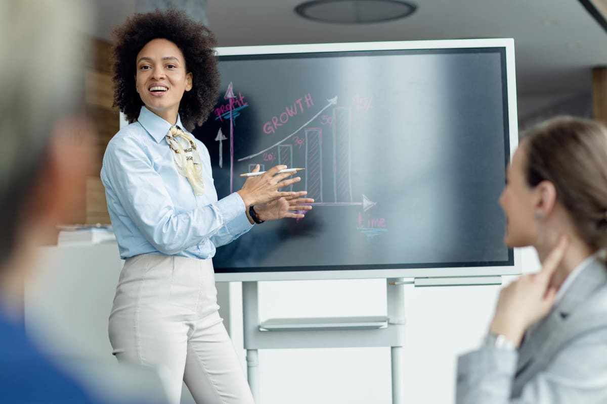 Reunião: importância do marketing pessoal Mulher em reunião de trabalho de pé na frente de seus colegas enquanto exibe dados em painel