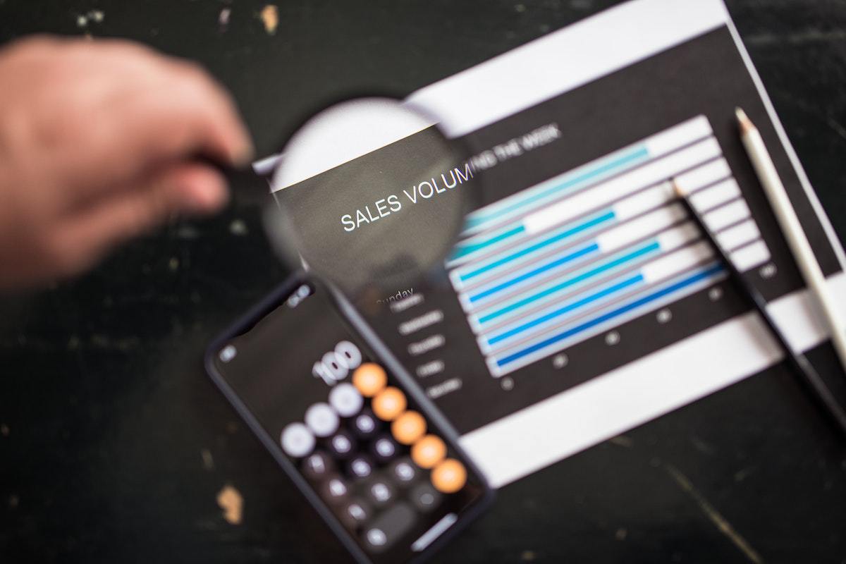 Homem com lupa sobre folha de papel e smartphone com foco na frase SALES VOLUM e objetos em desfoco no fundo da foto