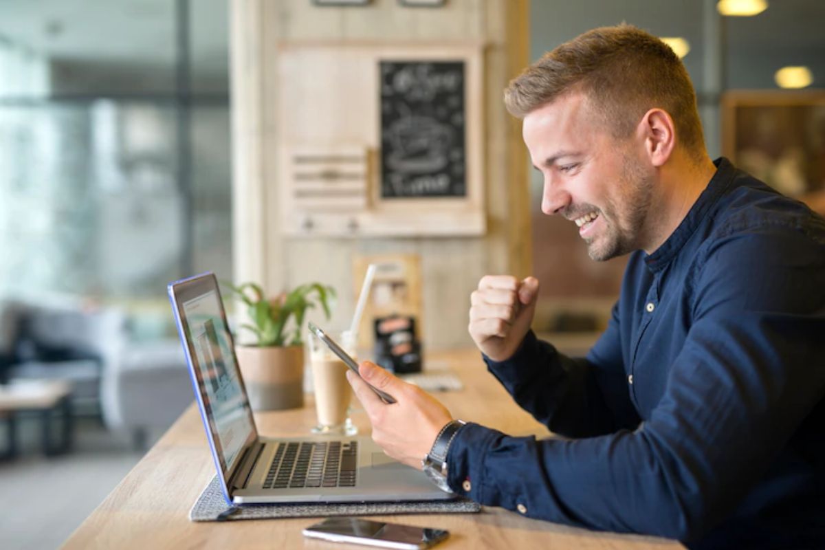 Homem sentado diante de mesa de madeira usando celular e notebook enquanto faz gesto de comemoração e sorri