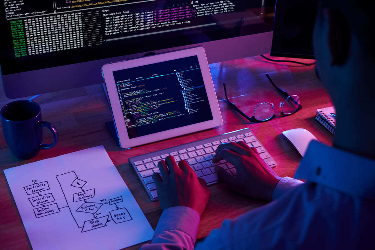 Pessoa programando em tablet na horizontal apoiado em suporte e com teclado sem fio, além de projeto desenhado em folha a4 à esquerda e monitor grande atrás do tablet, além de outros objetos na mesa
