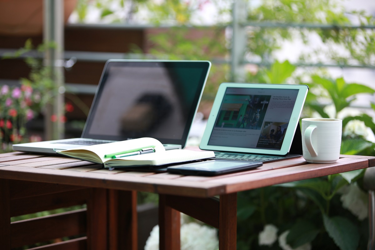 Notebook sobre mesa de madeira pequena ao lado de tablet de pé na horizontal, diante de agenda aberta que apoia canela e do lado de celular e caneca branca