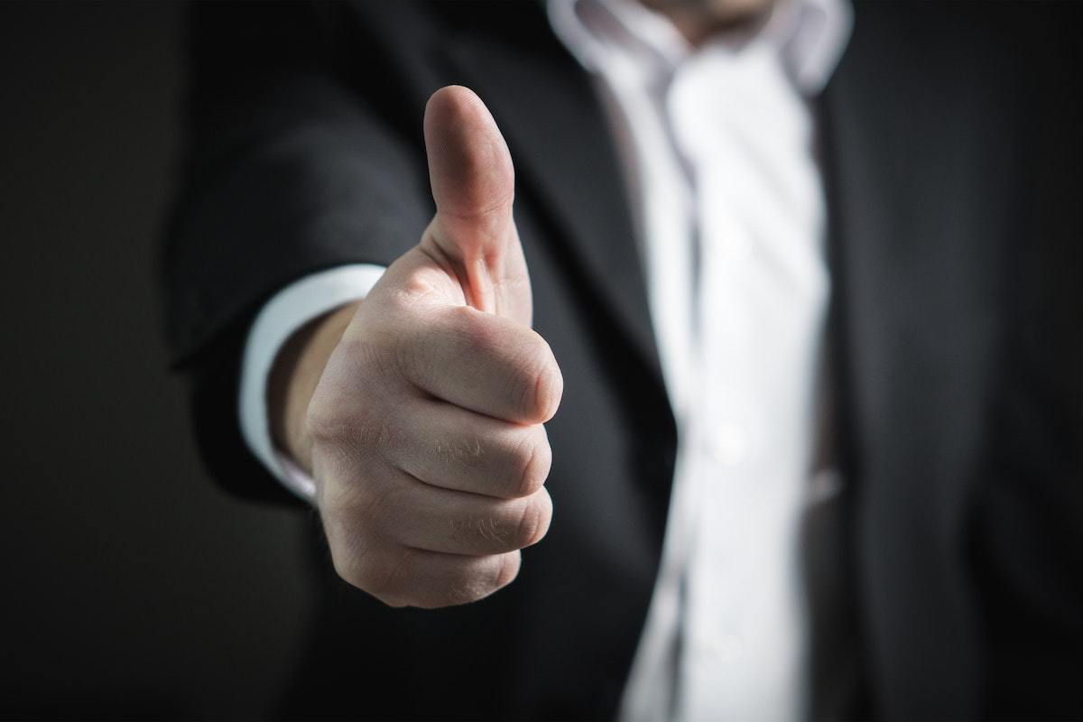 Homem com roupa social composta de camisa e blazer preto com braço direito esticado fazendo sinal de positivo com a mão, com seu polegar erguido