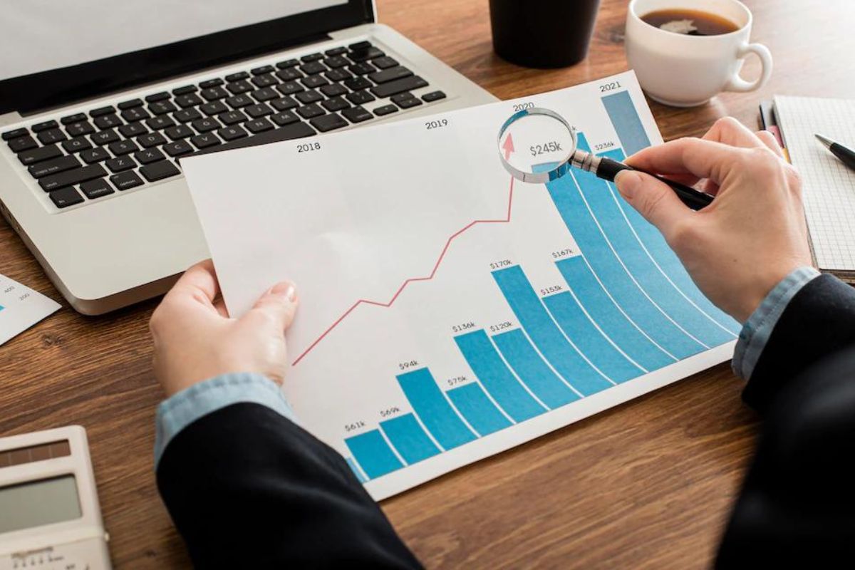 Gráficos: Marketing de resultados Pessoa sentada diante de mesa de madeira com notebook à frente analisando dados com lupa em folhas impressas com gráficos azuis