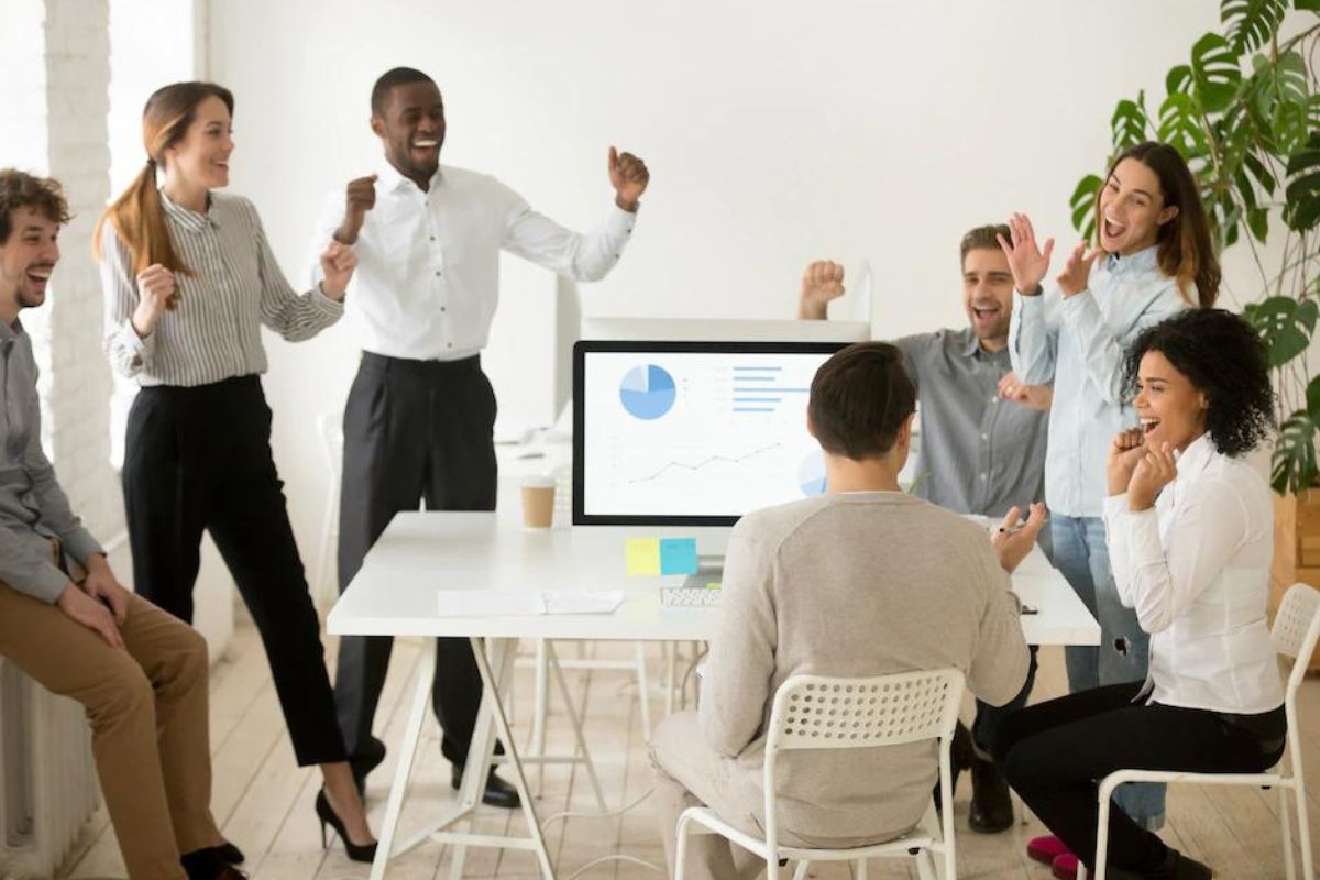 Reunião: Marketing de resultados Pessoas reunidas em torno de mesa branca quadrada, alguns sentados e outros em pé, em reunião com monitor no centro da mesa e comemorando sorridentes