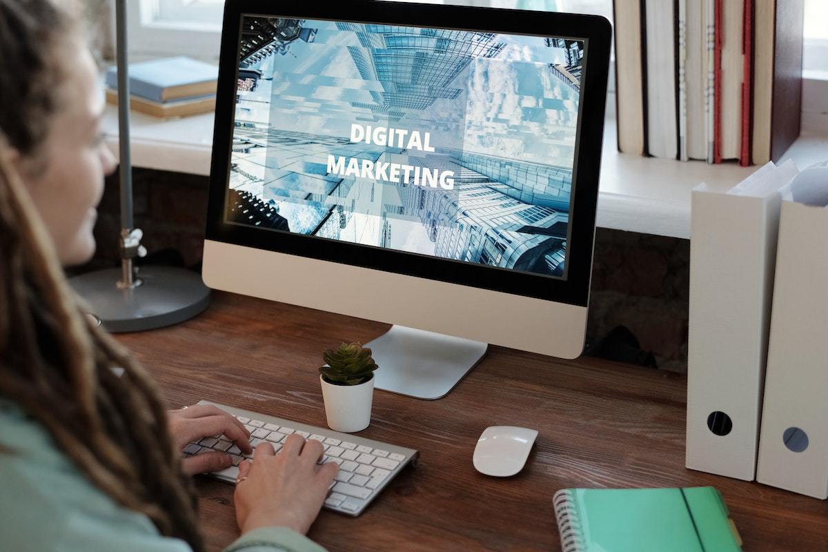 Computador: Marketing digital para iniciantes Mulher sentada diante de computador sobre mesa de madeira com fichários à direita e agenda diante de janela de vidro em escritório