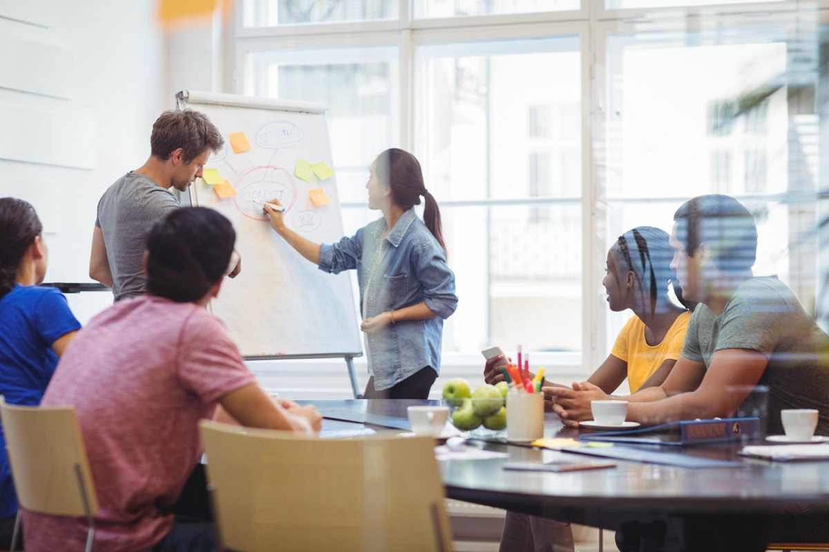 Reunião de trabalho com homens e mulheres sentados ao redor de mesa retangular com outro casal de pé analisando e explicando dados em painel de papel com post-its e dados colados no mesmo com janela de edifício de vidro ao fundo da imagem em desfoco