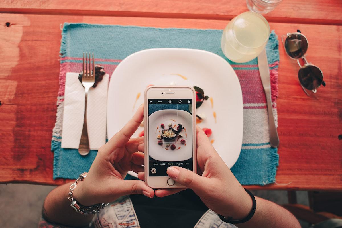 Mãos de mulher tirando foto de comida em restaurante com prato sobre jogo americano listrado com talheres e copo ao redor, além de óculos de sol
