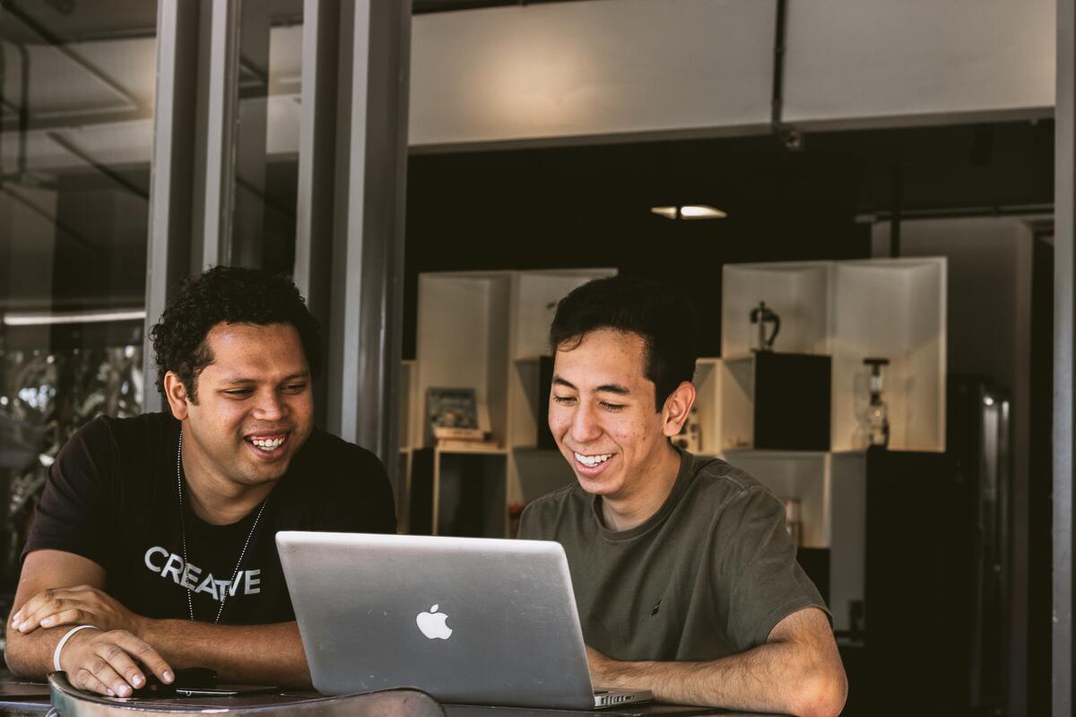 Dois homens sentados diante de notebook da Apple sorridentes enquanto analisa, algo em ambiente interno de empresa