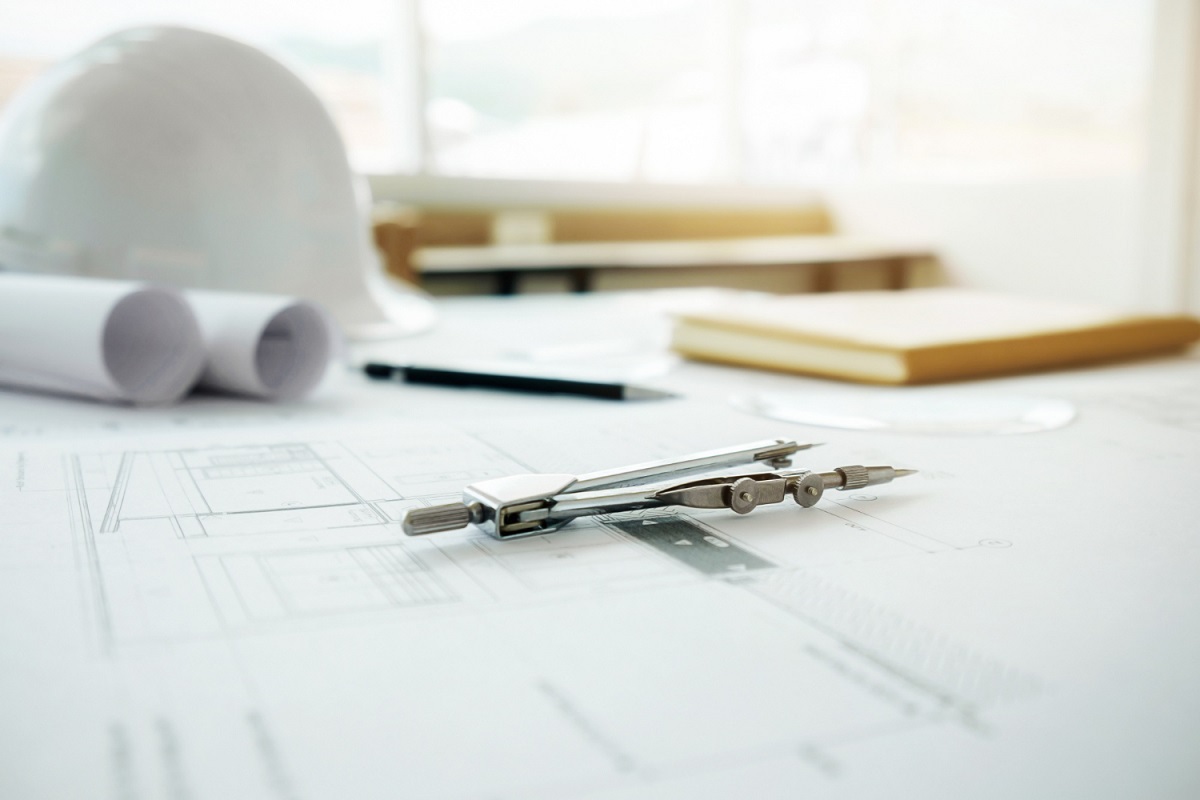 Compasso: Marketing na Arquitetura Compasso em foco sobre projeto ou planta feita por arquiteto em mesa grande com folhas enroladas à esquerda do objeto, além de lápis de madeira e cadernos em desfoco mais ao fundo