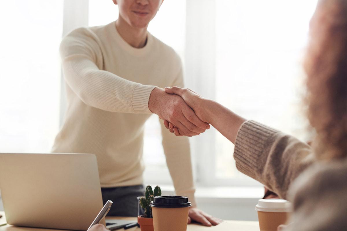 Acordo: Marketing para empresas de tecnologia Homem de pé em escritório cumprimenta mulher sentada do outro lado da mesa em que ele está diante, com notebook e copo de café em cima da mesma