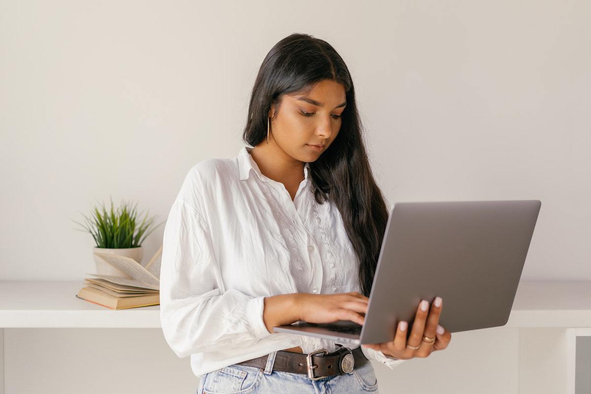 Notebook: Marketing para empresas de tecnologia Mulher de pé em ambiente de interior com planta decorativa ao fundo e unhas grandes segurando notebook cinza nas mãos e concentrada, com cabelo sobre o ombro jogado pra frente