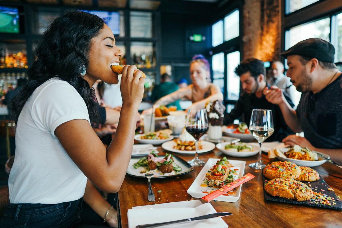 Pessoas comendo: marketing para restaurantes Pessoas sentadas ao redor de mesa de madeira em restaurante comendo refeição e sobremesa de grandes cookies em bandeja sobre a mesa