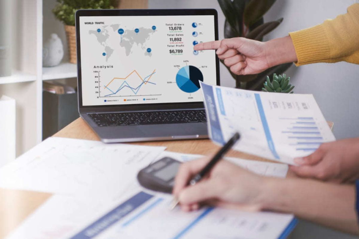 Pessoa analisando métricas em folha impressa com outra por trás apontando dados em tela de monitor à direita da imagem