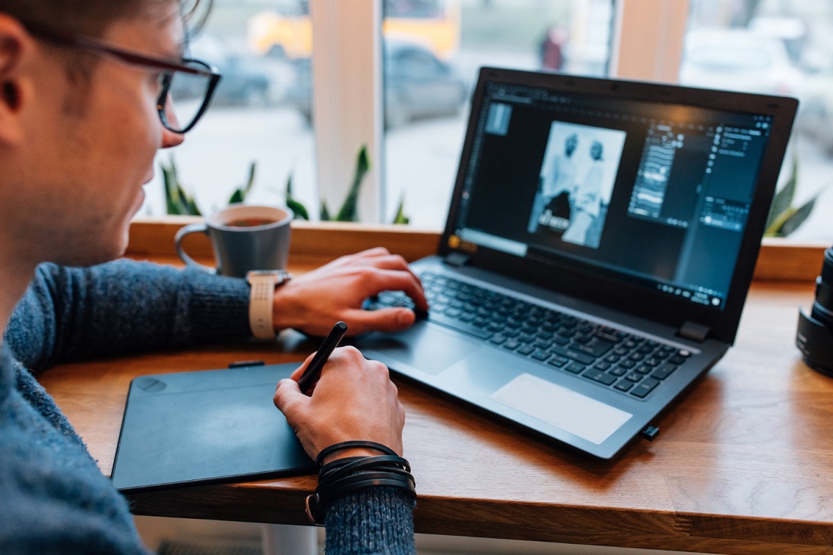 Homem sentado diante de mesa de madeira usando notebook e desenhando algo em aparelho digital que transforma tudo em desenho de Material design