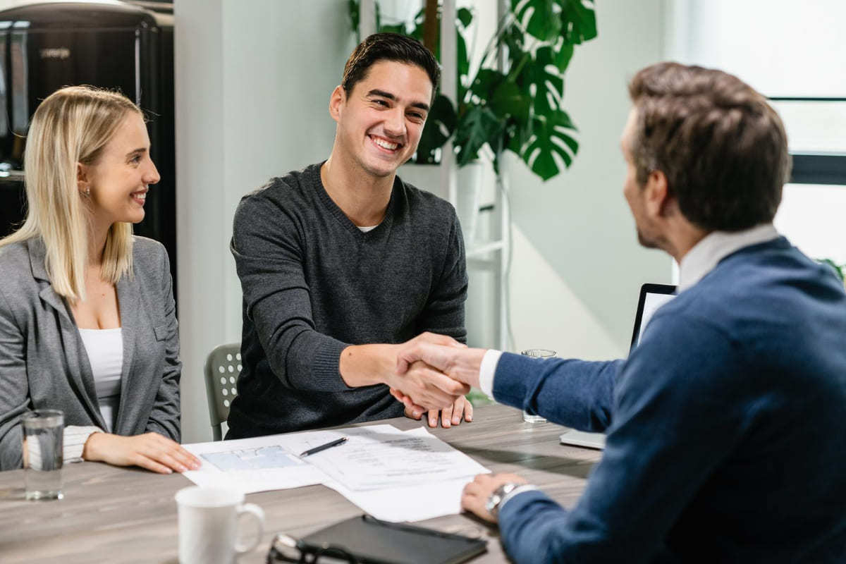 Pessoal sorrindo: onboarding Homem de paletó diante de mesa de escritório apertando mão de homem do outro lado da mesa e mulher ao seu lado sorri