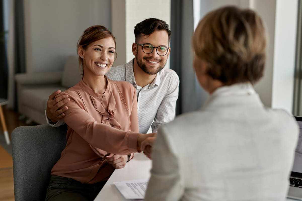 Mulher apertando mão de casal que está sorridente em local análogo à escritório