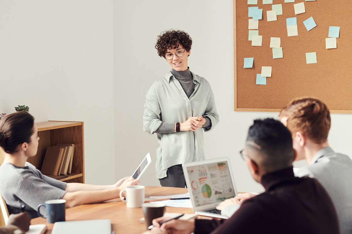 Local de trabalho: palestra motivacional Pessoas em local de trabalho com mulher de cabelo curto de pé diante de três companheiros e ao lado de painel com post-its enquanto fala sobre assuntos de trabalho