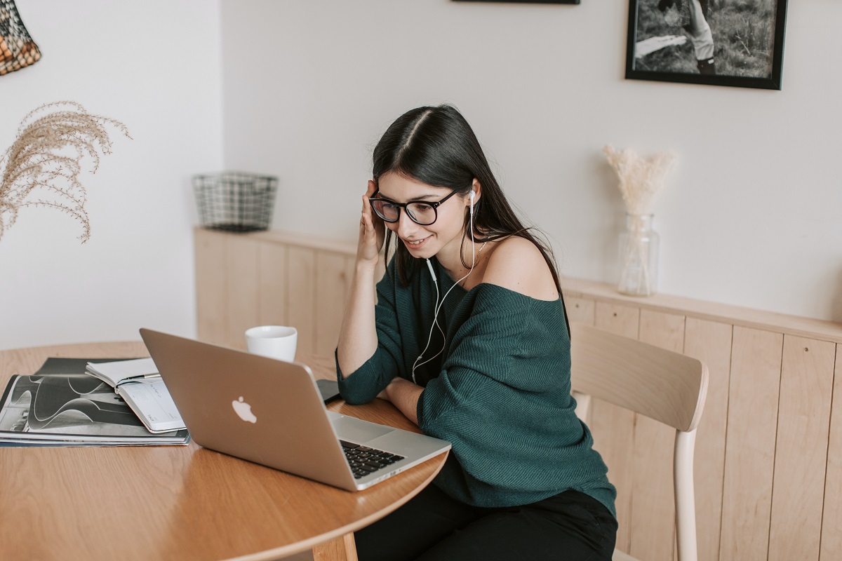 Mulher em local de trabalho sentada em cadeira de madeira diante de MacBook enquanto assiste à palestra motivacional no aparelho