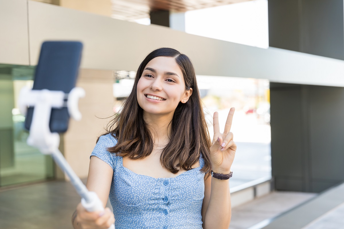 Mulher sorri e faz sinal de V com os dedos enquanto tira foto em celular com ajuda de pau de selfie