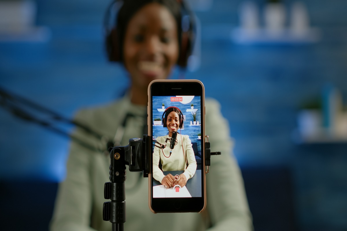 Celular em pedestal enquanto grava vídeo de mulher com fone de ouvido estilo headphone e fundo azul com alguns objetos na parede