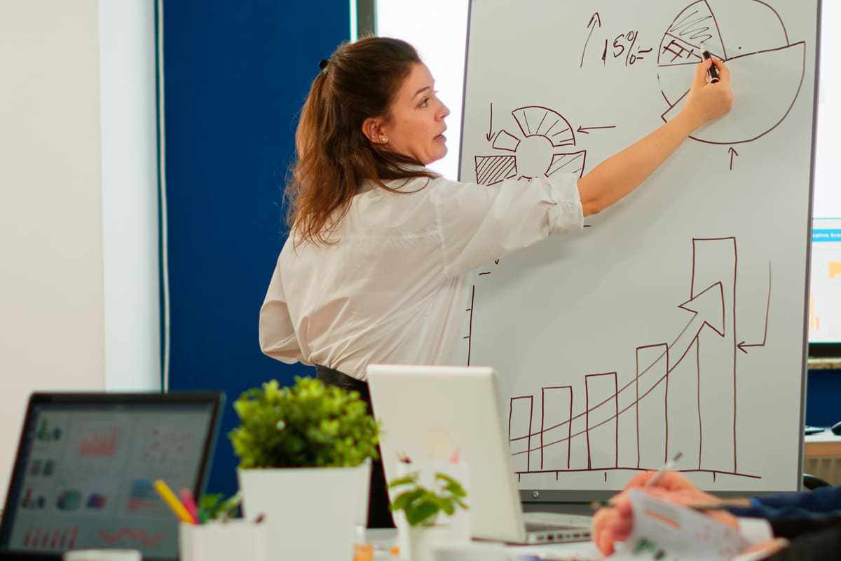Mulher explicando gráficos em painel para indivíduos em reunião de trabalho cujo seus notebooks aparecem na imagem