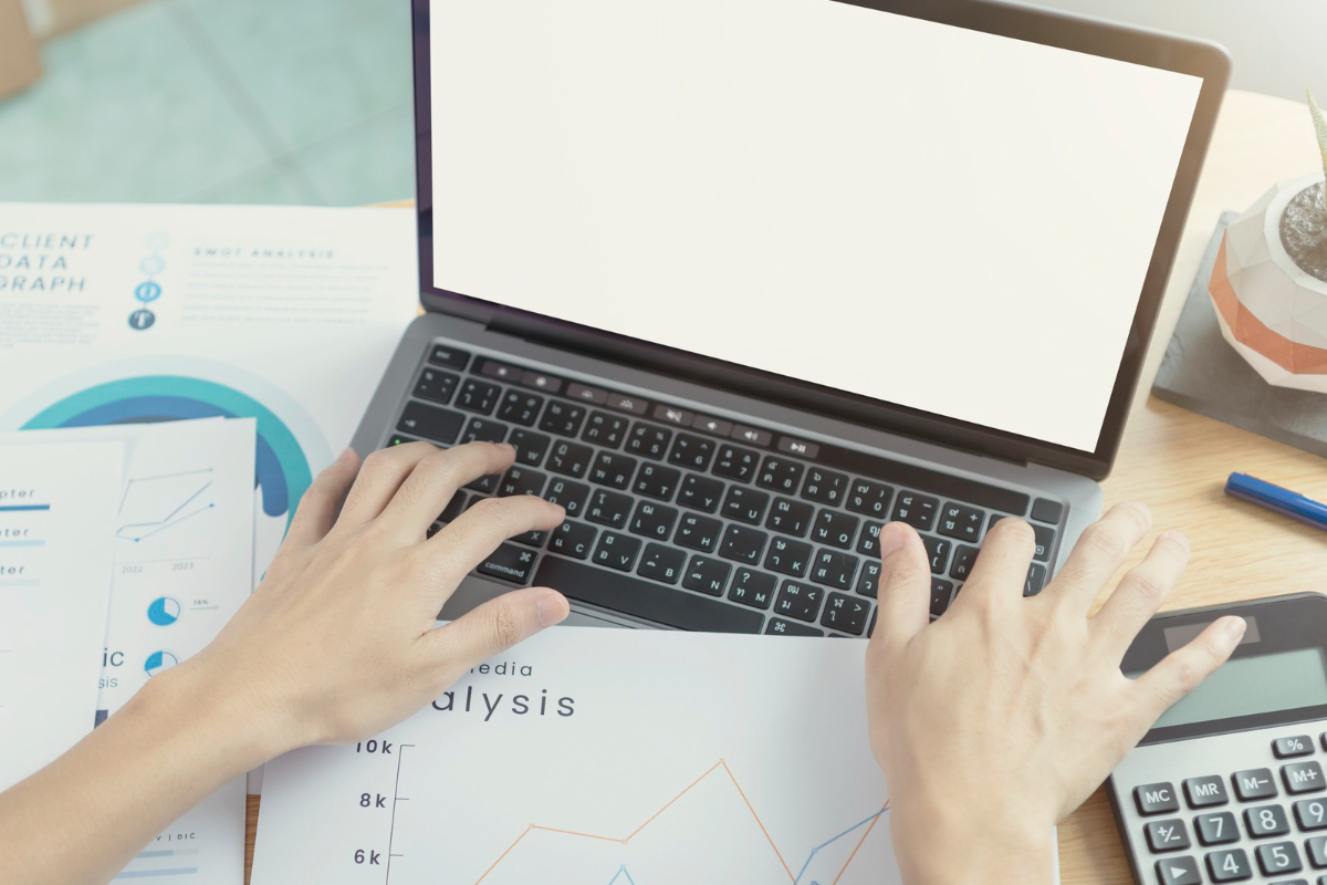 Pessoa digitando: Press release Pessoa digitando algo em notebook com folhas com gráficos impressos cobrindo parte do teclado