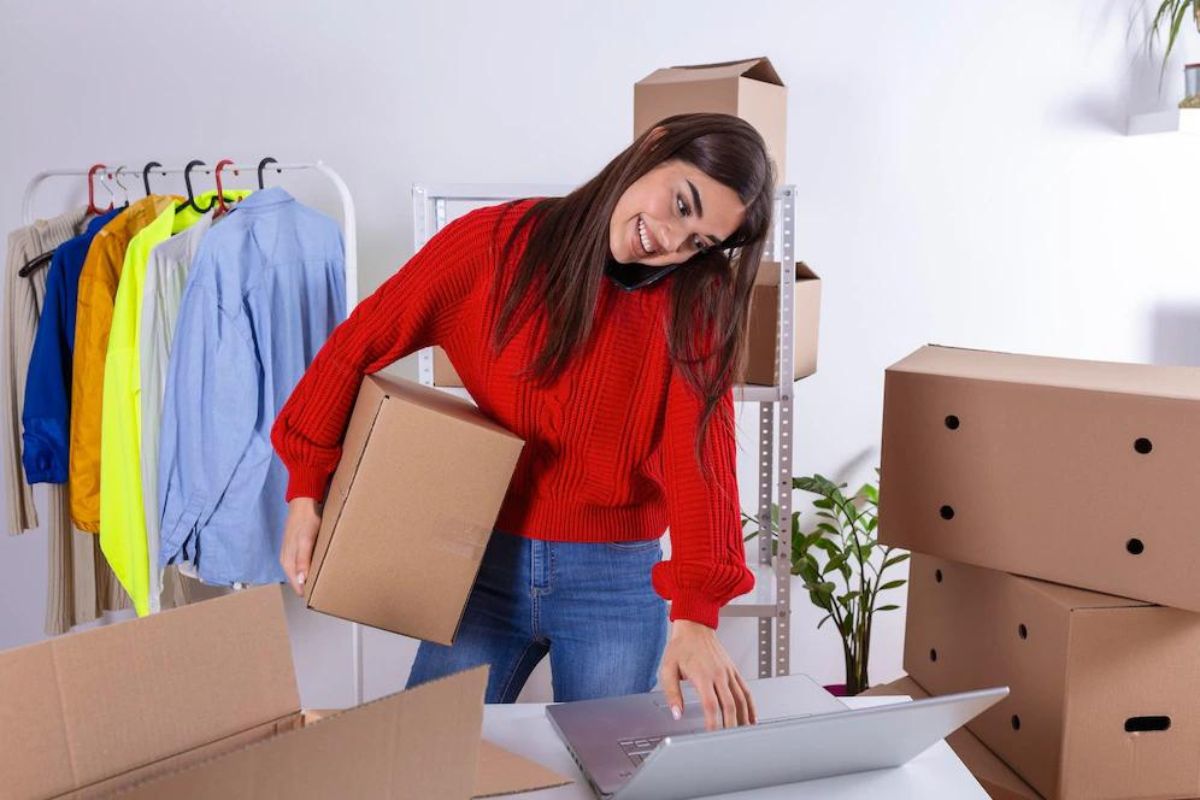 Mulher segurando caixas: profissões mais procuradas Mulher de calça jeans e suéter vermelho segurando caixa com a mão direita, usando notebook que está sobre mesa com a outra mão e algumas caixas espalhadas ao redor da sala e com roupas atrás da mesma