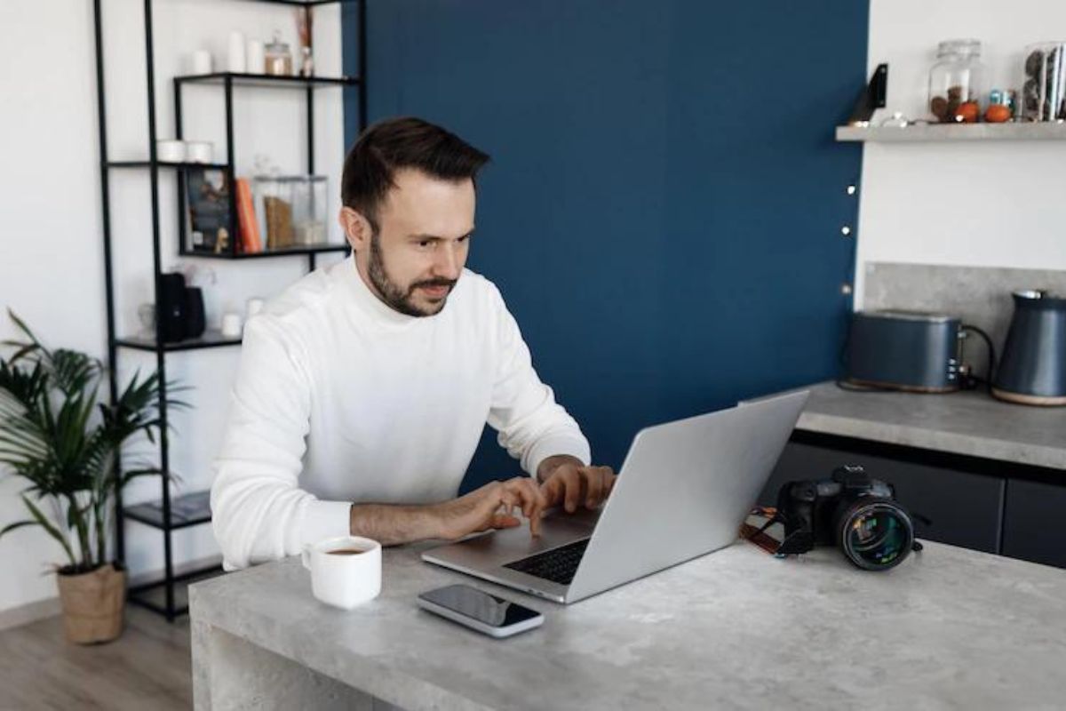 Homem com camisa de mangas compridas branca trabalhando em notebook sobre mesa cinza com celular ao lado, além de caneca branca e câmera, com estante com objetos ao fundo e planta de interior