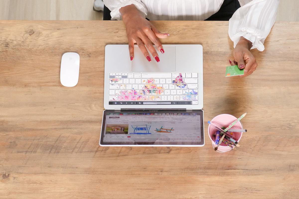 Mulher usando notebook sobre mesa de madeira com unhas longas e pintadas de rosa segurando cartão de crédito com uma das mãos e com porta-lápis ao lado do PC