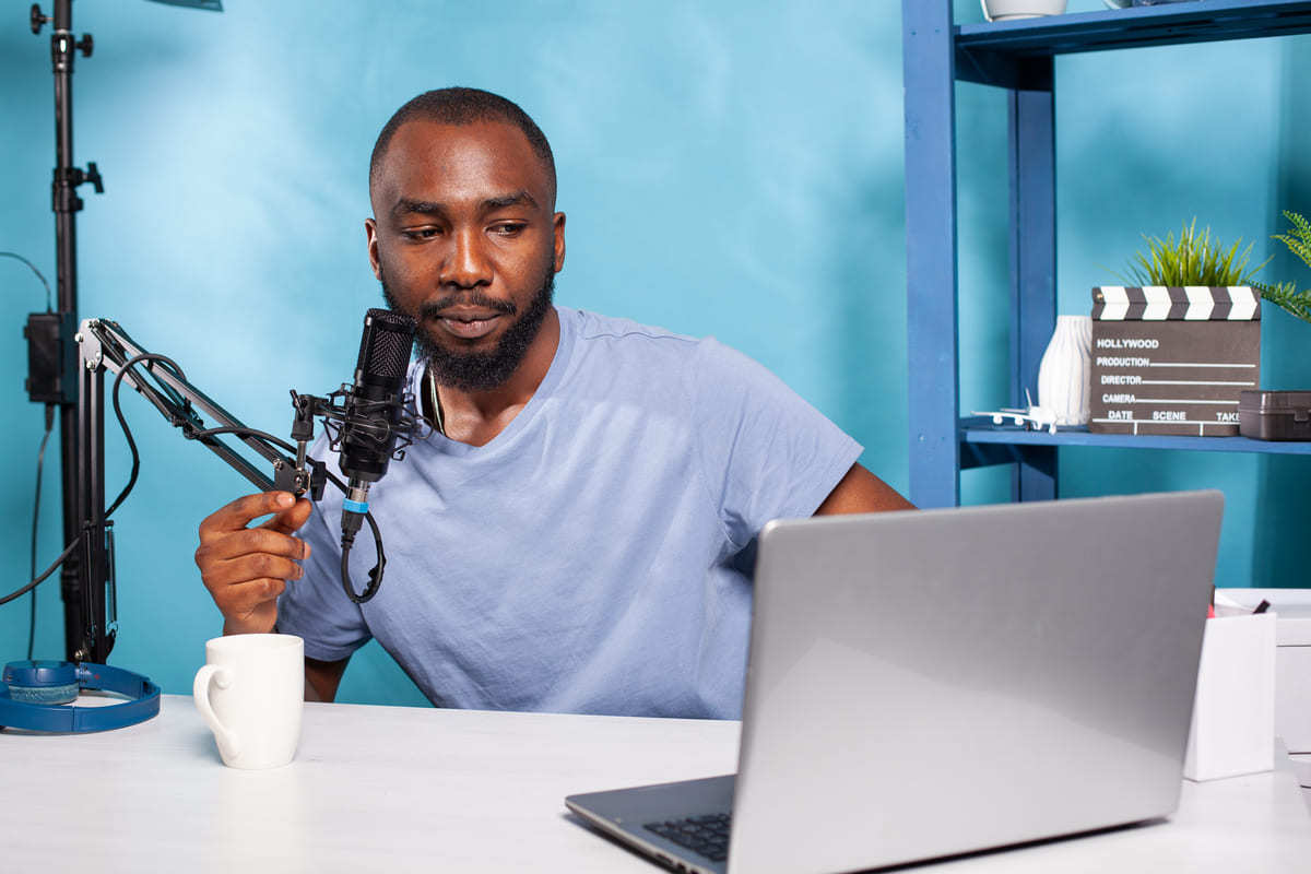 Homem falando algo em microfone de podcast em pedestal diante de mesa branca que suporta caneca e notebook cinza com parede ao fundo azul e estante de mesma cor à direita da foto com objetos diversos, como claquete