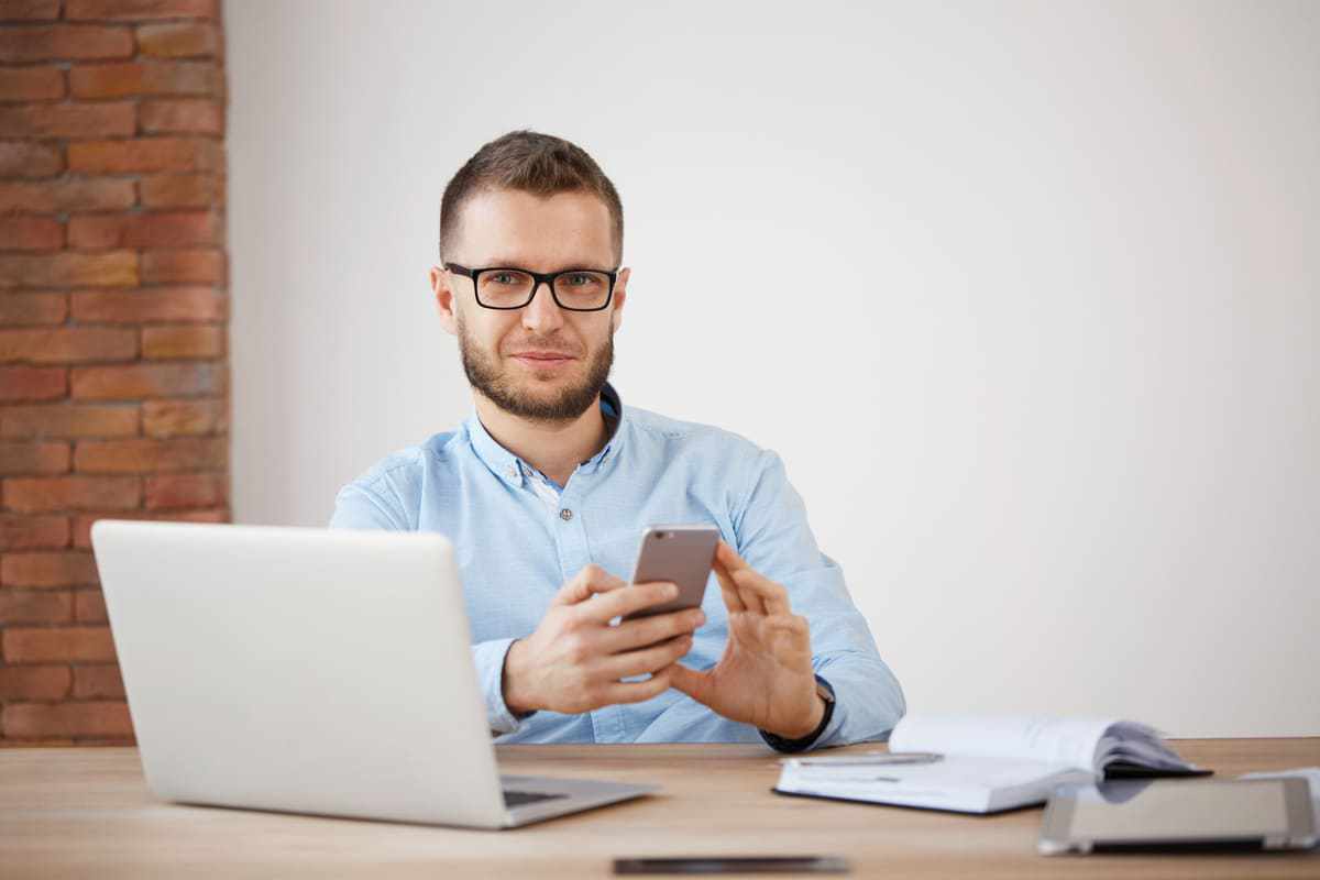 Homem com ar sorridente sentado diante de mesa de madeira com agenda, tablet e notebook e segurando celular