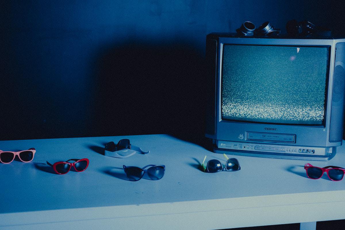 Televisão de tubo antiga sobre mesa de madeira com óculos de sol estilo retrô espalhados sobre a mesma e com pouca luz na foto