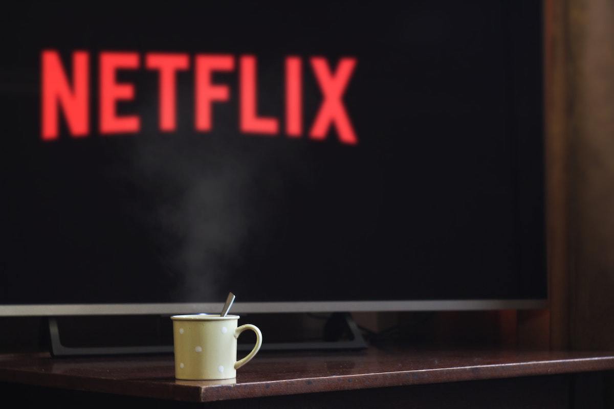 Televisão grande em tela de entrada da Netflix em móvel de madeira com caneca branca na frente da mesma com colher de metal pequena dentro da mesma
