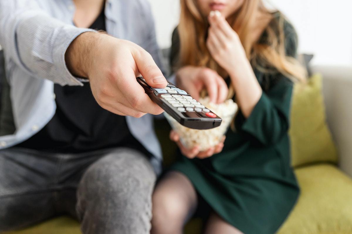 Homem e mulher sentados em sofá e comendo pipoca em balde próprio para tal enquanto o homem aponta controle remoto de TV para frente