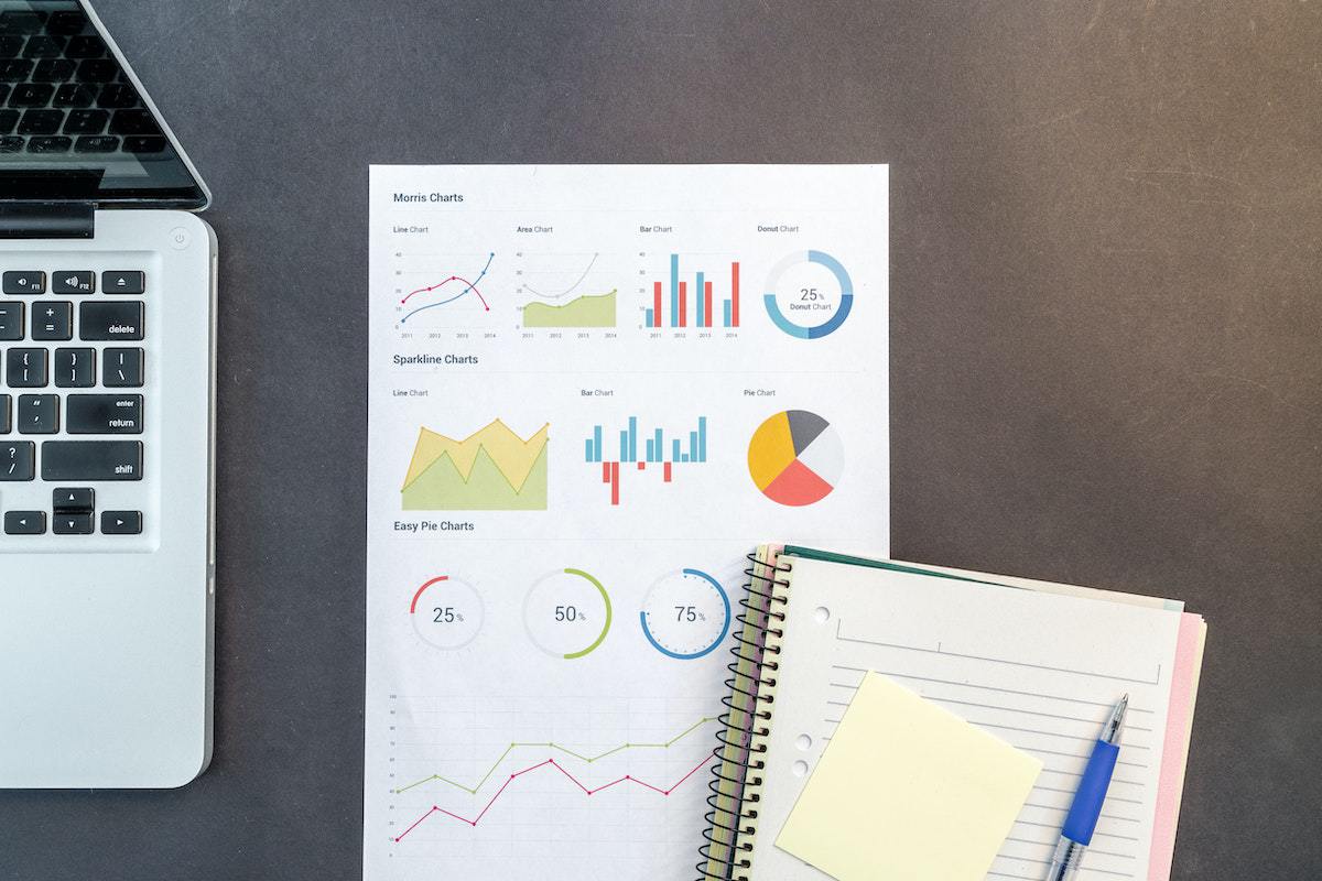 Papel com gráficos impressos embaixo de agenda com post-its e uma caneta azul sobre mesa de cor cinza e ao lado de um notebook cinza
