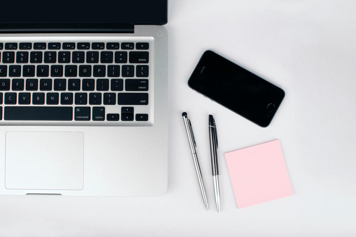 Notebook cinza com tela desligada, mas aberto, em cima de mesa branca ao lado de duas canetas e bloco de post-it com rosa, além de smartphone preto mais à frente