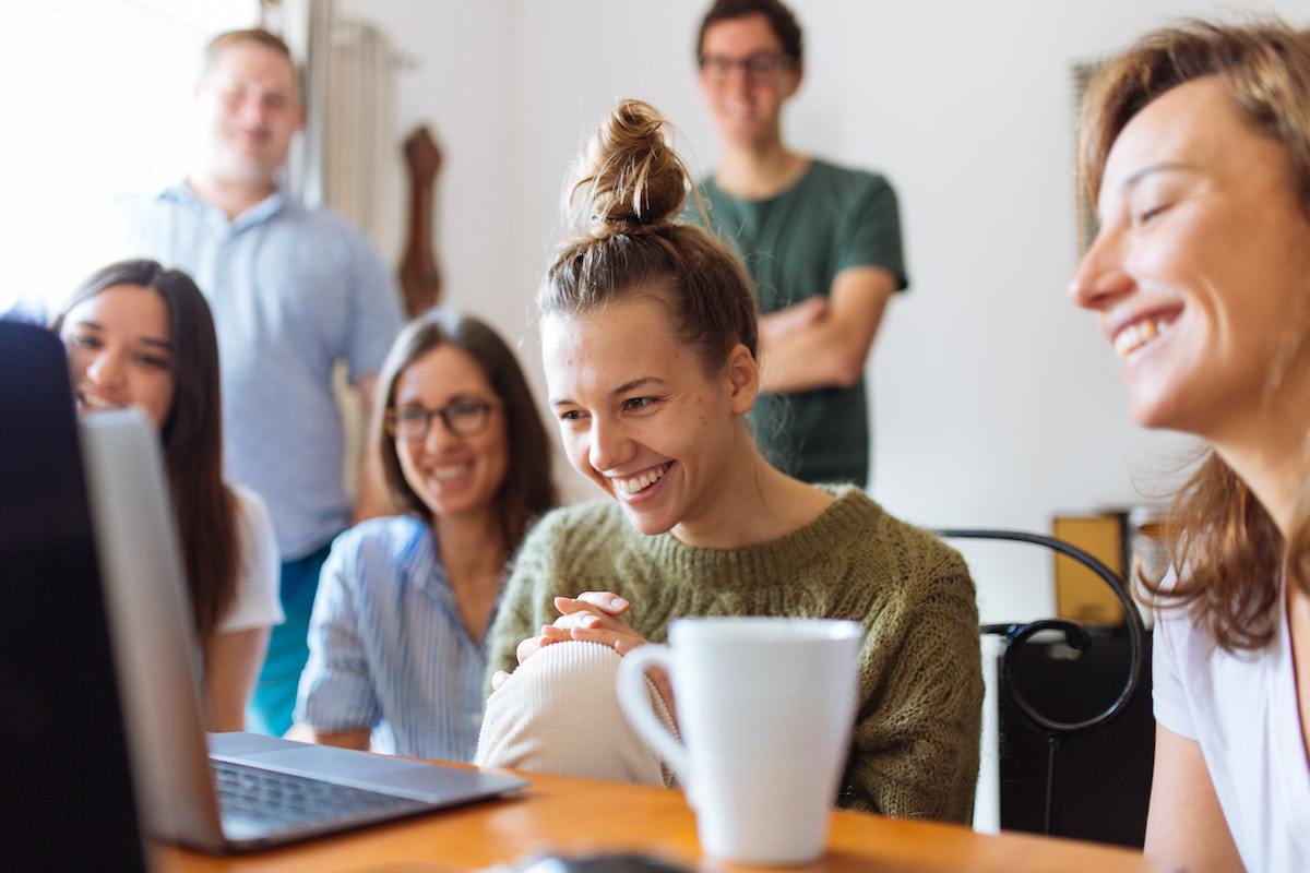Local de trabalho: Tipos de vídeo Quatro mulheres sentadas diante de mesa de madeira analisando algo em notebook com caneca à esquerda e dois homens de pé em local de trabalho