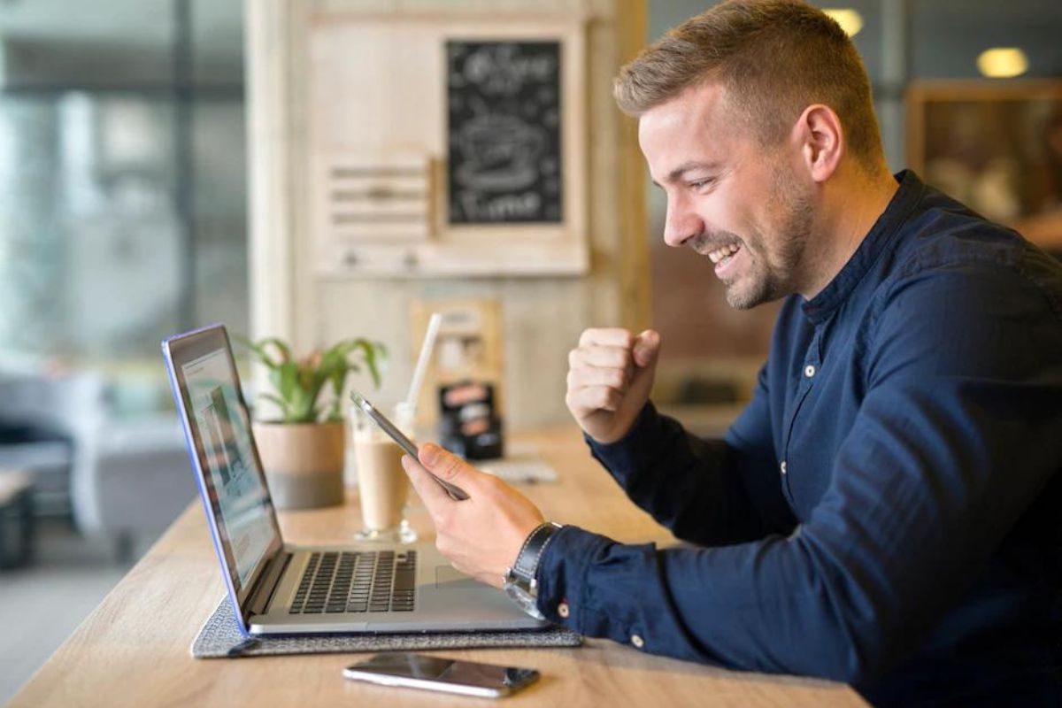 Celular: Unir marketing e vendas Homem sentado diante de balcão de madeira usando notebook e celular e comemorando com gesto de mão enquanto sorri