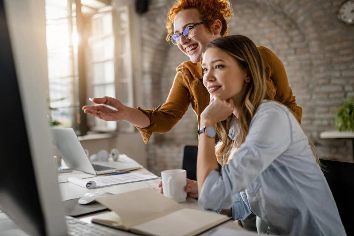 Computador: Unir marketing e vendas Duas mulheres, uma sentada e uma em pé, conversando enquanto sorriem e apontam para algo em computador sobre mesa branca